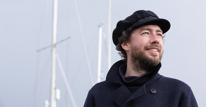 Historian Oskar Cox Jensen in a coat and hat smiling with boat masts in the background